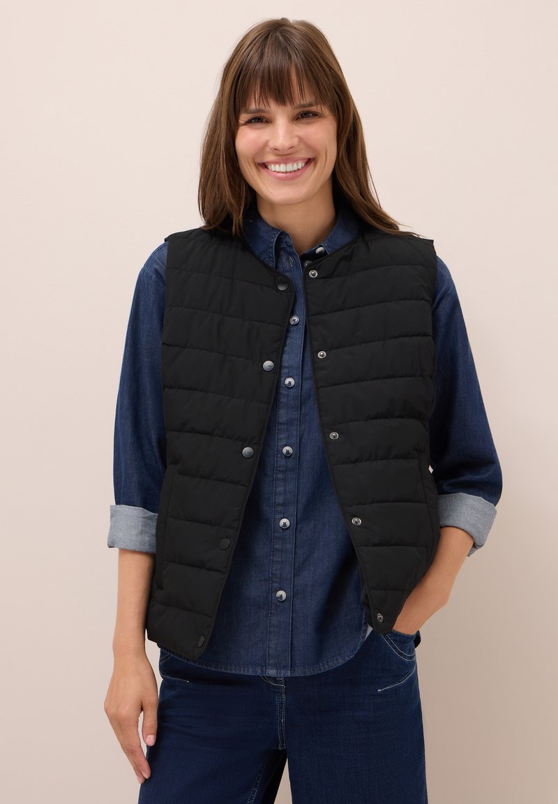 Donna sorridente che indossa un gilet trapuntato nero sopra una camicia di denim azzurro con le maniche arrotolate e jeans blu scuro.