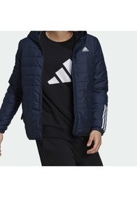 Osoba nosící námořnicky modrou prošívanou bundu Adidas přes černou mikinu Adidas s velkým bílým logem, oříznutou u krku a boků.