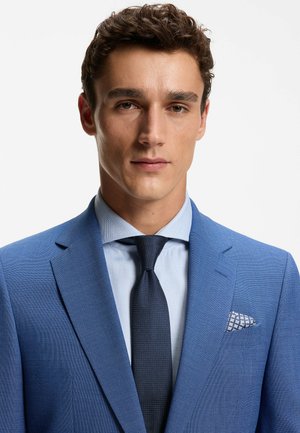 Nuori mies, jolla on kiharat ruskeat hiukset, pukeutunut vaaleansiniseen paitaan, tummansiniseen teksturoituun solmioon ja kirkkaan siniseen pukuun, jossa on kuvioitu taskuliina.