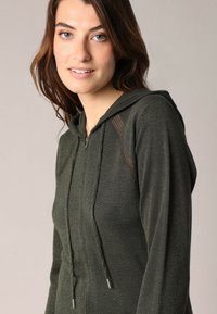 Donkergroene rits-vest met een gestructureerde brei, een kap met trekkoord en mesh zijkanten. Heeft een aansluitende pasvorm en zachte stof.