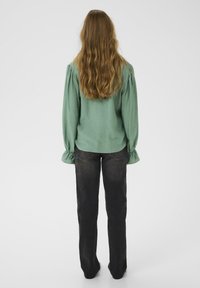 Blusa verde menta con maniche a sbuffo, abbinata a jeans di denim scuro, vista da dietro. Tessuto morbido e vestibilità rilassata.