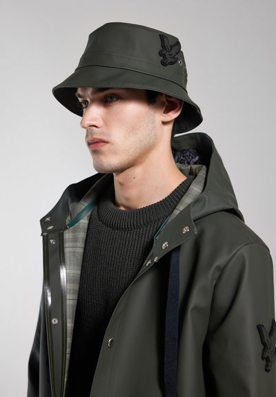 Grüner wasserdichter Bucket Hat mit Adleremblem; kombiniert mit einem grünen Regenmantel mit kariertem Futter und einem strukturierten Strickpullover darunter.