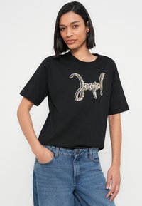 Μαύρο μπλουζάκι με μεγάλο διακοσμητικό κείμενο σε μεταλλικό χρυσό χρώμα. Το μπλουζάκι έχει κοντά μανίκια και άνετη γραμμή, συνδυασμένο με μπλε τζιν.