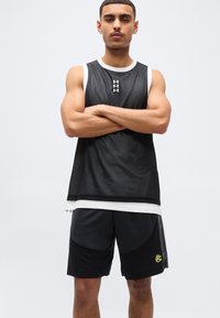 Νέος άνδρας με σταυρωμένα χέρια, φορώντας μαύρο αμάνικο τοπ από δίχτυ πάνω από λευκό πουκάμισο και μαύρο αθλητικό σορτς με κίτρινο λογότυπο, στέκεται μπροστά σε απλό φόντο.