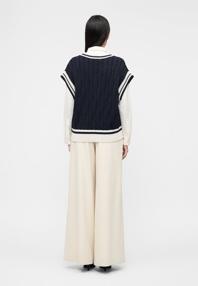 Femme aux longs cheveux noirs portant un pull sans manches en maille torsadée bleu marine par-dessus une chemise blanche à manches longues et un pantalon large crème, vue de dos.