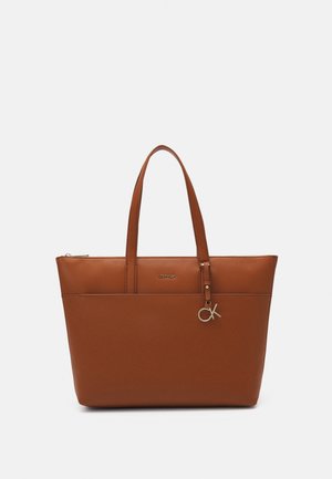 Bolso tote de cuero marrón con dos asas, cierre de cremallera y un pequeño charm metálico con las letras "CK" colgando cerca del asa.