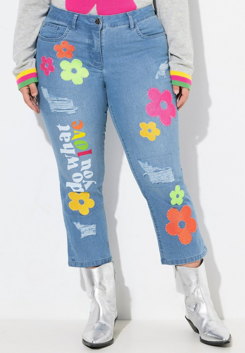 Jean en denim bleu clair avec des appliqués floraux aux couleurs vives, des détails déchirés et un texte brodé "fais ce que tu aimes" sur la jambe.