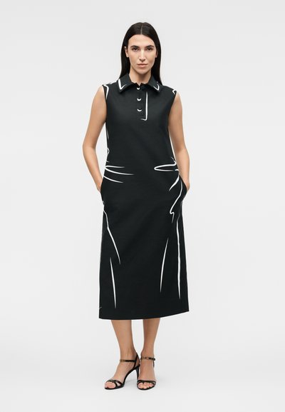 Γυναίκα που φοράει μαύρο μίντι φόρεμα χωρίς μανίκια με λευκές αφηρημένες γραμμές, γιακά, κουμπιά και μαύρα πέδιλα με λουριά, στέκεται με τα χέρια στις τσέπες.