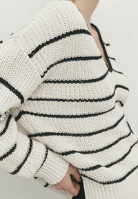 Pull en maille avec une base blanche et des rayures noires horizontales. Il présente un col en V et des manches amples avec des poignets côtelés.