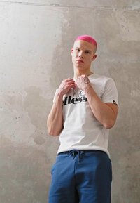 Hvit t-skjorte med svart logo, kombinert med blå joggebukser. Huden er lys, håret er rosa. Individet er i en nøytral setting med betongvegger.
