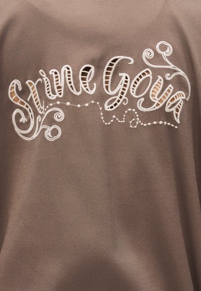 Καφέ ύφασμα με λευκή κεντητή καλλιγραφική γραφή "Suivre Gaïa" και διακοσμητικές σβούρες και τελείες.