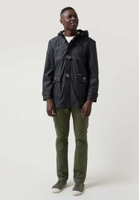 Chaqueta negra impermeable con capucha, con cierres de toggles dorados, dos bolsillos frontales y un parche de logo azul, llevada sobre pantalones verde oliva.