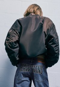 Crna satenska bomber jakna s nabranim rukavima, uparena s plavim trapericama koje imaju natpise "HAWKINS" i "STRANGER THINGS".