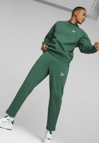 Grüner Pullover und passende Jogginghosen aus weichem Stoff. Mit dezentem Logo auf der Brust und dem Hosenbein. Getragen mit weißen Sportschuhen.
