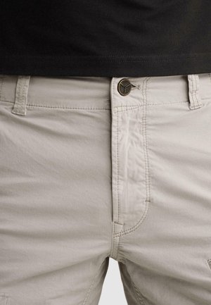 Gros plan d'un pantalon beige avec une taille boutonnée et des coutures apparentes, porté avec une chemise noire rentrée.