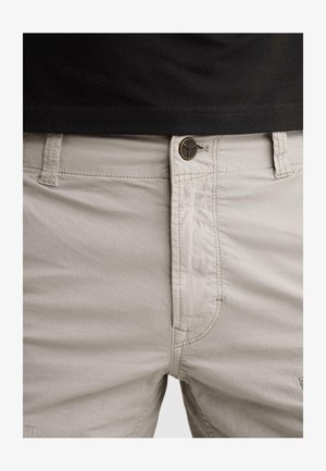 Detail béžových kalhot s knoflíkovým pasem a viditelným prošíváním, nošených s vtaženou černou košilí.