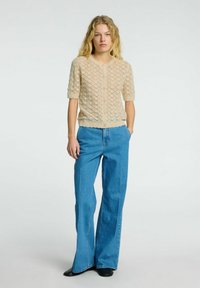 Beige pletený kardigan s krátkými rukávy, se vzorem v diamantovém stylu a zapínáním na knoflíky, spárovaný s modrými kalhotami s širokými nohavicemi.