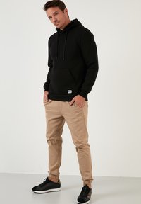 Sudadera negra con capucha y bolsillo delantero, combinada con pantalones beige ajustados y zapatillas negras con un patrón multicolor en el costado.