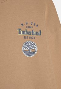 Il tessuto di cotone color sabbia presenta un logo circolare blu di Timberland con un albero, completato da testo blu e giallo che indica il marchio e la località.