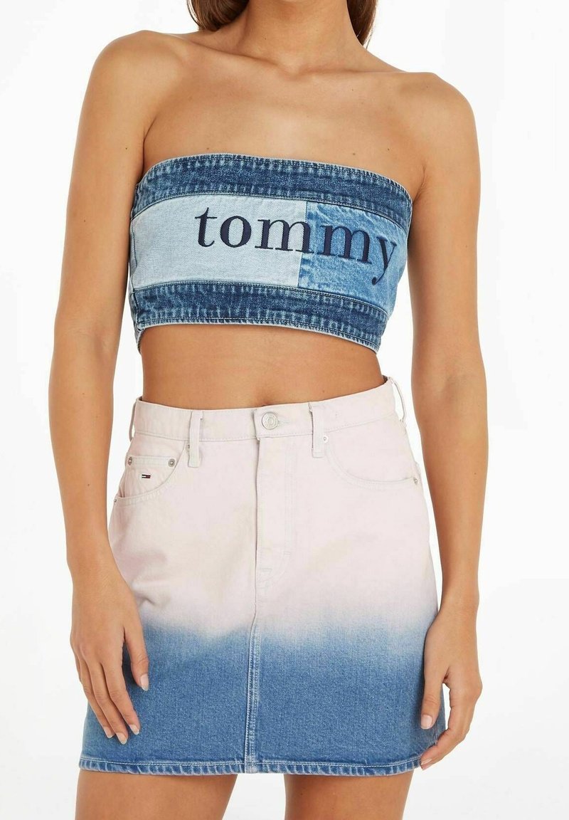 Femme portant un crop top en denim sans bretelles avec le texte "tommy" et une jupe en denim taille haute dégradée du blanc au bleu.