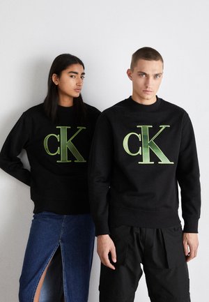 Sudaderas negras con un gran logo verde "CK". La tela parece suave y texturizada. Los modelos llevan pantalones negros y de mezclilla, posando frente a un fondo sencillo.