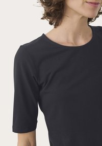 Frau, die ein schlichtes schwarzes T-Shirt mit kurzen Ärmeln und einem runden Halsausschnitt trägt, was ihren Hals, ihre Schulter und einen Teil ihres Gesichts zeigt.