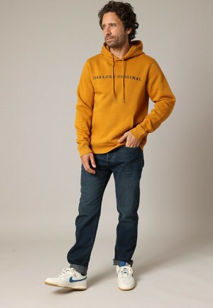 Haut à capuche jaune moutarde avec cordons de serrage et poche avant, associé à un jean bleu foncé et des baskets blanches avec des accents bleus.