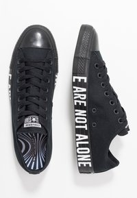 Baskets en toile noire avec un bout en caoutchouc noir brillant, portant une inscription blanche sur le côté disant « NOUS NE SOMMES PAS SEULS ». Matières textile et caoutchouc.