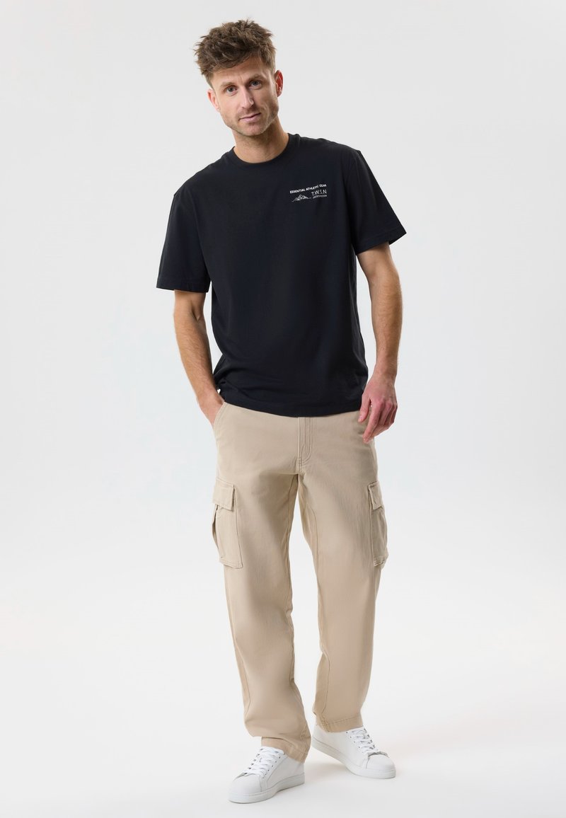 Homme debout, mains dans les poches d'un pantalon cargo beige, portant un T-shirt noir avec un texte blanc et des baskets blanches, sur un fond uni.