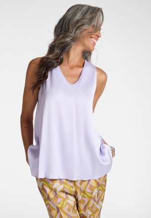 Kvinde med langt gråt hår, der smiler skævt, iført en ærmeløs lavendel-top og mønstrede bukser i gul, pink og grøn.