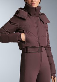 Donker bordeauxrode pufferjas met een hoge kraag, ritssluitingen op de zakken en een taille met riem. Voorzien van een zachte, met bont gevoerde capuchon en een doorgestikte textuur.