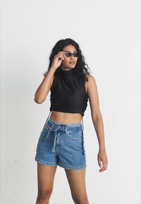 Zwarte mouwloze crop top met gathered zijkanten, gecombineerd met hoge blauwe denim shorts. Het model draagt een donkere zonnebril tegen een neutrale achtergrond.