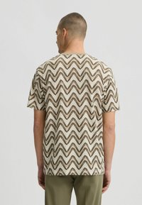 Camiseta de manga corta en color crema con patrón de zigzag marrón. Cuello redondeado, corte relajado, hecha de tela ligera.