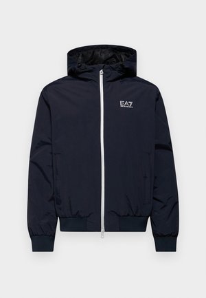 Giacca windbreaker blu navy con cappuccio, chiusura frontale con zip, polsini elastici e logo EA7 sul petto. Materiale liscio e leggero.
