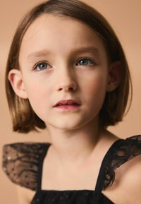 Robe noire avec des manches transparentes et volantes, présentant une texture et un éclat subtils. Un fond pâle complète les traits doux d'une jeune fille.
