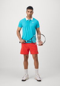 Jasnoniebieska koszulka polo z białym kołnierzykiem, pomarańczowe szorty i rakieta tenisowa. Białe skarpetki sportowe i kolorowe buty sportowe dopełniają outfit.