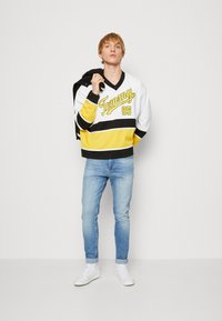 Pullover in stile hockey di colore bianco con strisce gialle e nere, con logo "Tommy". Indossato con jeans azzurri chiari e sneakers bianche.