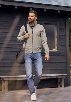 Un homme portant une veste grise à fermeture éclair et un jean marche devant une cabane en bois noire en portant un sac à bandoulière noir.
