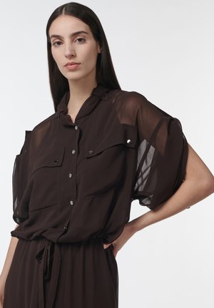 Mujer con cabello largo y oscuro, vestida con una blusa negra transparente con botones, bolsillos y cintura anudada, de pie con una mano en la cadera.