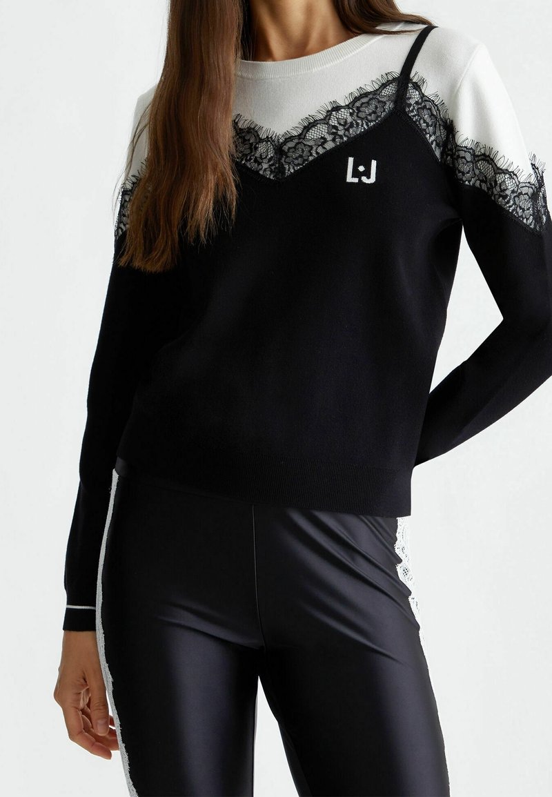 Pull tricot noir et blanc avec bordure en dentelle, manches longues et col rond, associé à des leggings noirs brillants avec des accents en dentelle.