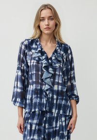 Blusa a scacchi blu e bianchi con colletto arricciato, maniche lunghe e vestibilità rilassata. Tessuto leggero e trasparente con una texture morbida e design a strati.