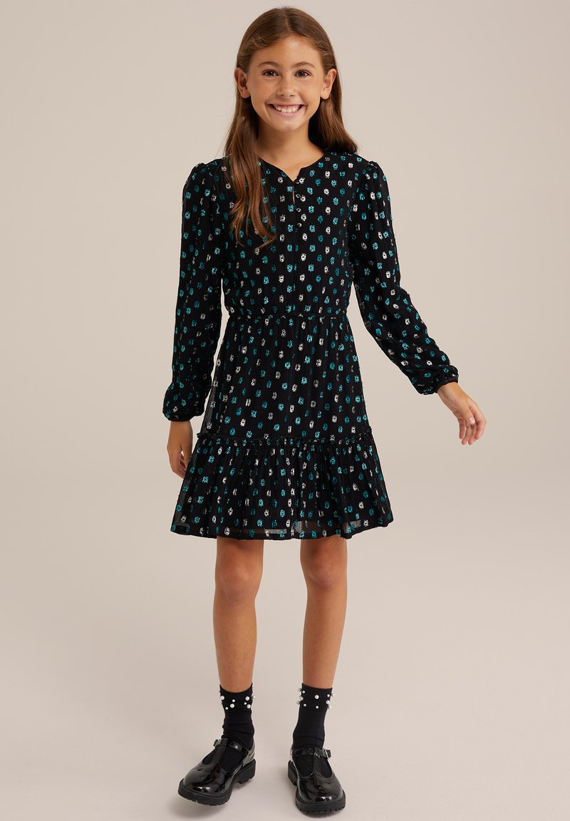 Schwarzes Kleid mit teal- und weißen Blumenmuster, langen Ärmeln, elastischem Bund, gerafftem Saum, kombiniert mit schwarzen Socken und Lackschuhen.