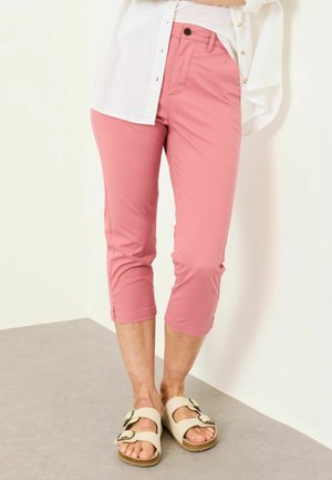 Personne portant un pantalon rose court et des sandales blanches à boucle, debout contre un mur et un sol clairs.
