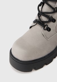 Bota de cuero gris en el frente con suela de goma negra gruesa y cordones negros atravesados por ojales metálicos, sobre fondo blanco.