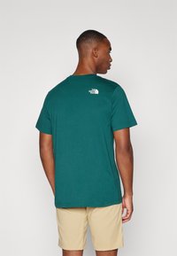Tricou din bumbac teal cu mâneci scurte, având un mic logo alb în partea superioară a spatelui. Purtați cu pantaloni scurți bej, prezentând un stil casual.