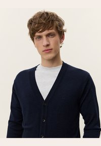 Cardigan navy in tessuto morbido, con scollo a V profondo, cinque bottoni e un discreto logo. Indossato sopra una camicia grigio chiaro.