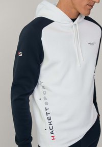 Mikina s kapucňou v bielej a námorníckej modrej farbe. Obsahuje predné vrecko, červené a čierne logo a mäkkú, textúrovanú tkaninu.