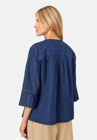 Top in denim blu scuro con scollatura rotonda, maniche 3/4 e dettaglio arricciato sul retro. Presenta applicazioni di bottoni sulle maniche e una vestibilità comoda.