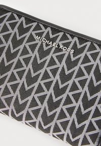 Portefeuille en tissu noir avec un motif zigzag blanc et le logo argenté "MICHAEL KORS". Texture lisse avec une forme rectangulaire élégante.