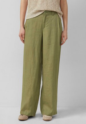 Osoba nosząca luźne lniane spodnie w oliwkowym kolorze z przodu zapinane na guziki oraz beżowy pleciony top, stojąca na szarej podłodze na tle jednolitej ściany.